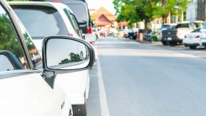 Zijspiegel van geparkeerde auto in smalle straat tijdens proefrit brommobiel met zicht op verkeer en parkeerruimte