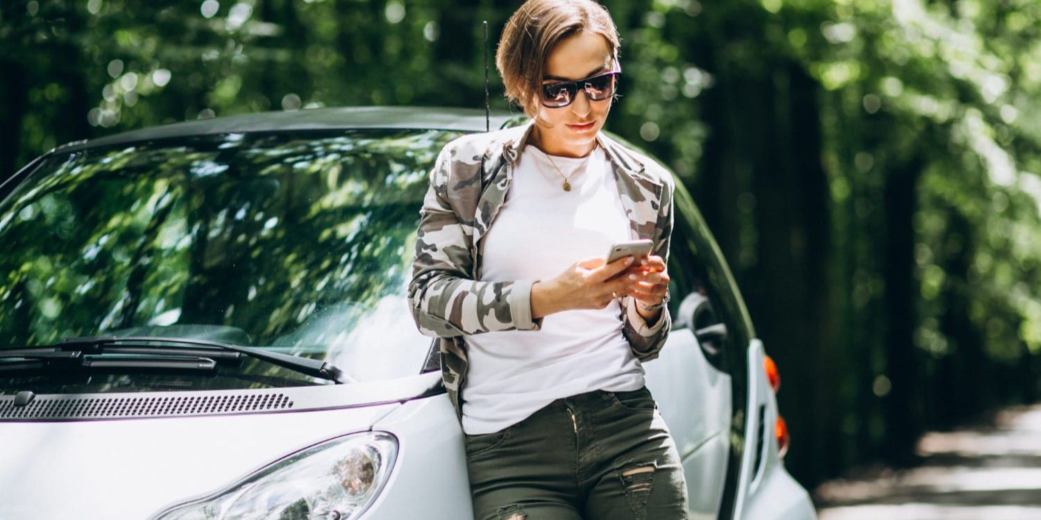 Persoon staat naast compacte auto in groene omgeving en bekijkt telefoon voor proefrit brommobiel
