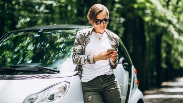 Persoon staat naast compacte auto in groene omgeving en bekijkt telefoon voor proefrit brommobiel