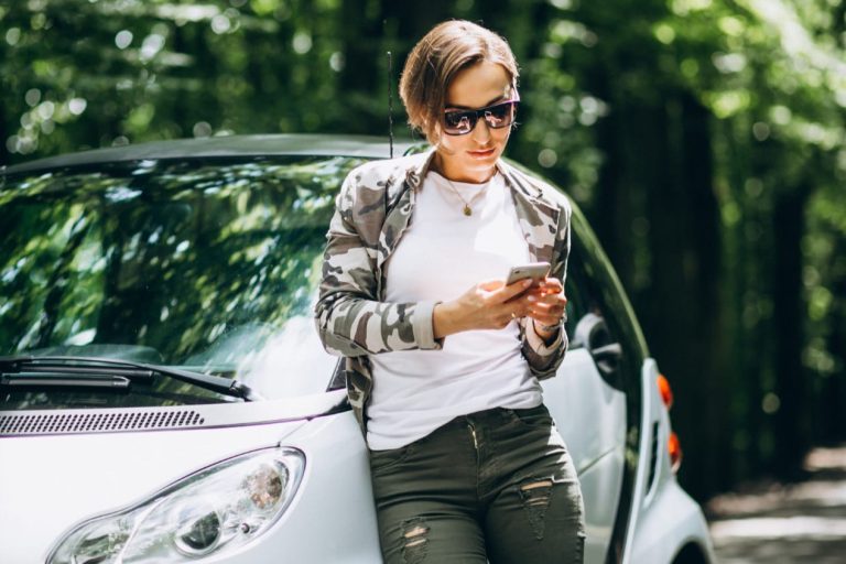 Persoon staat naast compacte auto in groene omgeving en bekijkt telefoon voor proefrit brommobiel