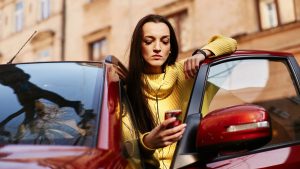 Bestuurder leunt tegen rode auto en bekijkt smartphone tijdens voorbereiding op proefrit brommobiel in de stad