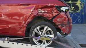 Close-up van een beschadigde rechterachterhoek van een rode auto, vastgemaakt met een spanband op een takelwagen. 